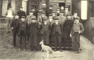 1908 Kangaroos team pictured in their street clothes standing in front of a fine house. The team mascot, a small kangaroo, is standing in front of the team.